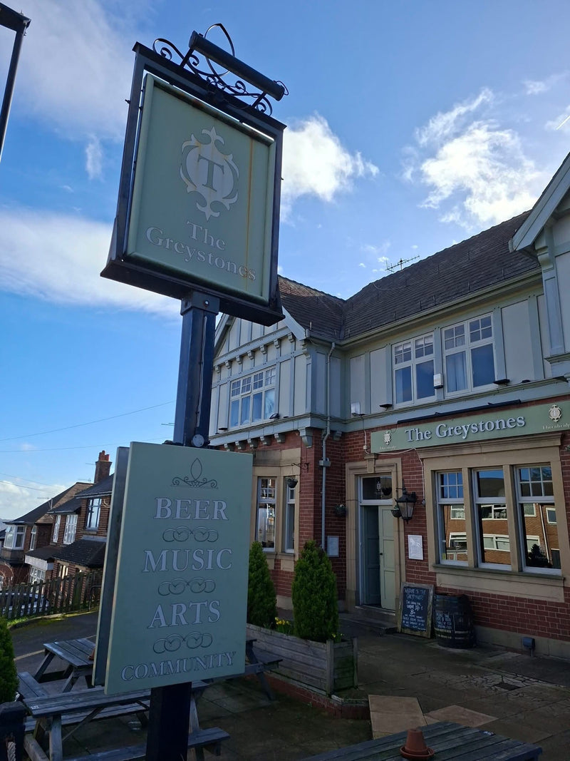 A pub exterior with a large hanging sign reading 'The Greystones' and a smaller sign advertising beer, music, arts, and comedy club. The building has brick and light-colored exterior walls, windows, and a blue sky with clouds in the background.