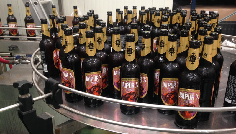 Multiple bottles of Jaipur X beer arranged on a conveyor belt in a brewery setting.