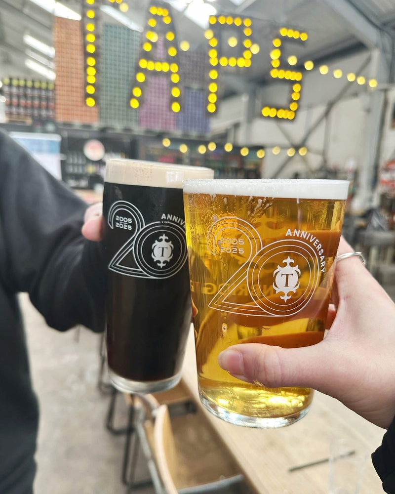 Two hands holding pint glasses with white 20-year anniversary logos, one filled with dark stout and one with golden lager, in a bar setting with a yellow 'TAPS' sign in the background.